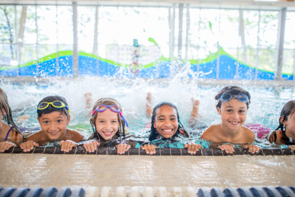 A multi-ethnic group of kids are taking a swimming class at an indoor pool. They are practicing kicking at the side of the pool, while smiling at the camera.